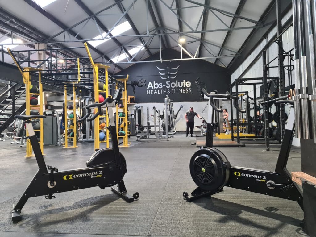 A spacious gym with a high ceiling and exposed beams. Two stationary bikes are in the foreground. Weightlifting equipment and a person working out are in the background. The wall displays the text "Abs-Solute Health & Fitness.