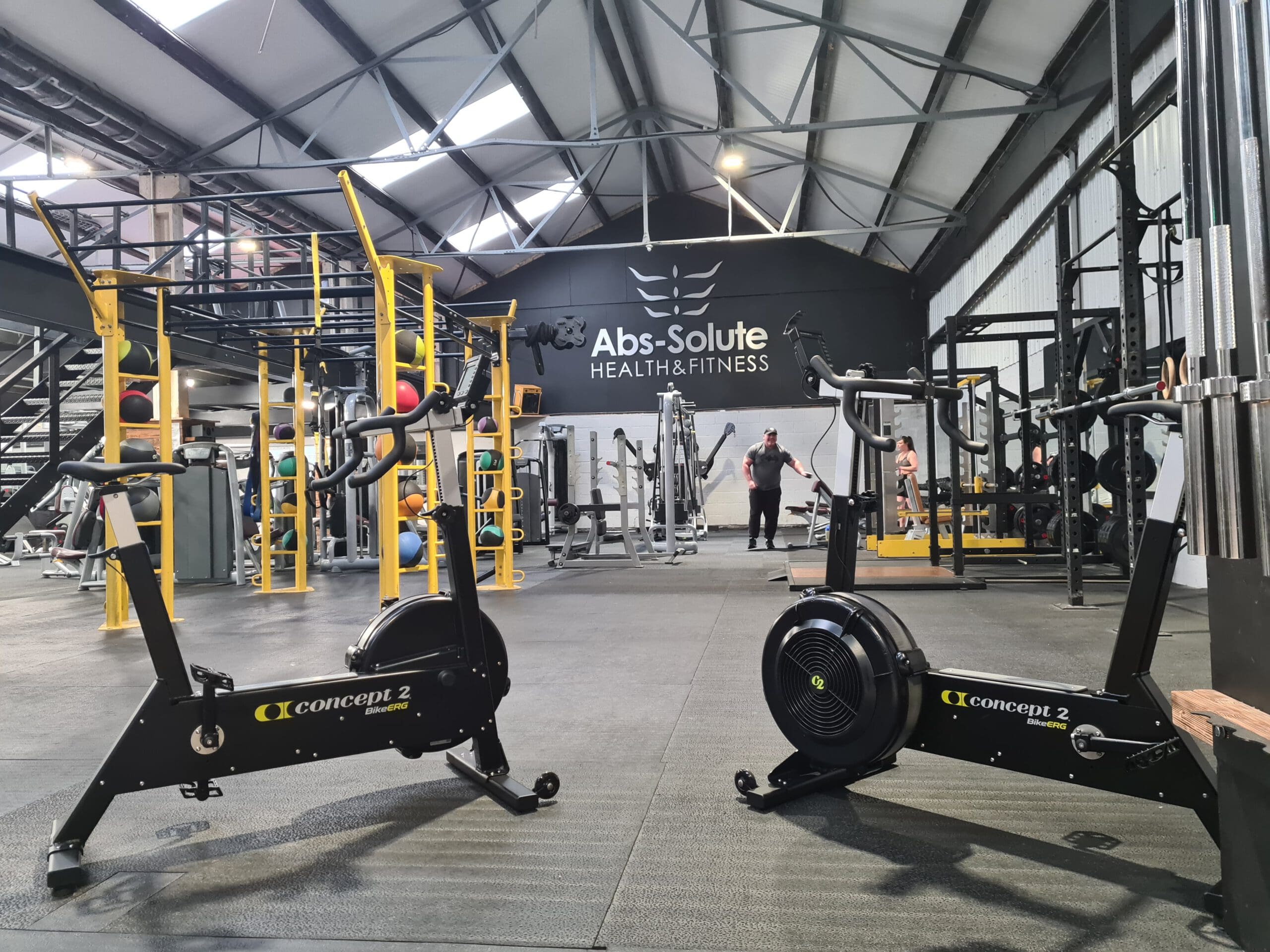A spacious gym with a high ceiling and exposed beams. Two stationary bikes are in the foreground. Weightlifting equipment and a person working out are in the background. The wall displays the text "Abs-Solute Health & Fitness.