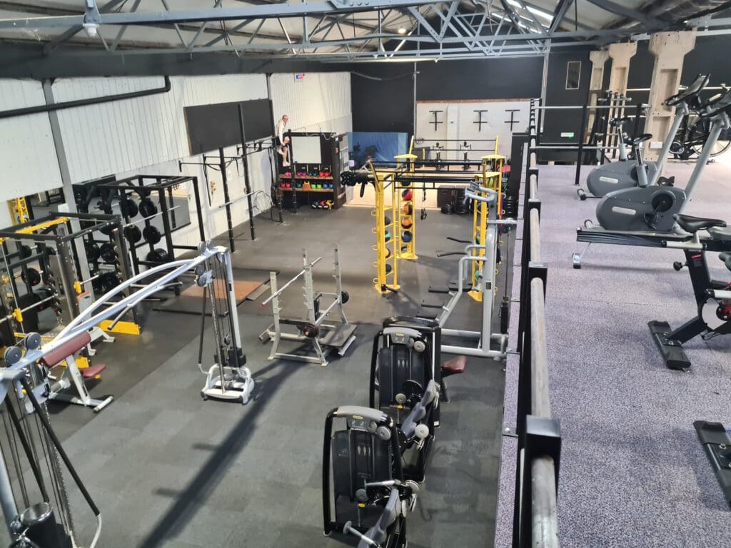 Wide view of a gym with various exercise equipment, including weight racks, machines, and stationary bikes. The space is organized and well-lit, with gray and yellow elements highlighting some equipment.