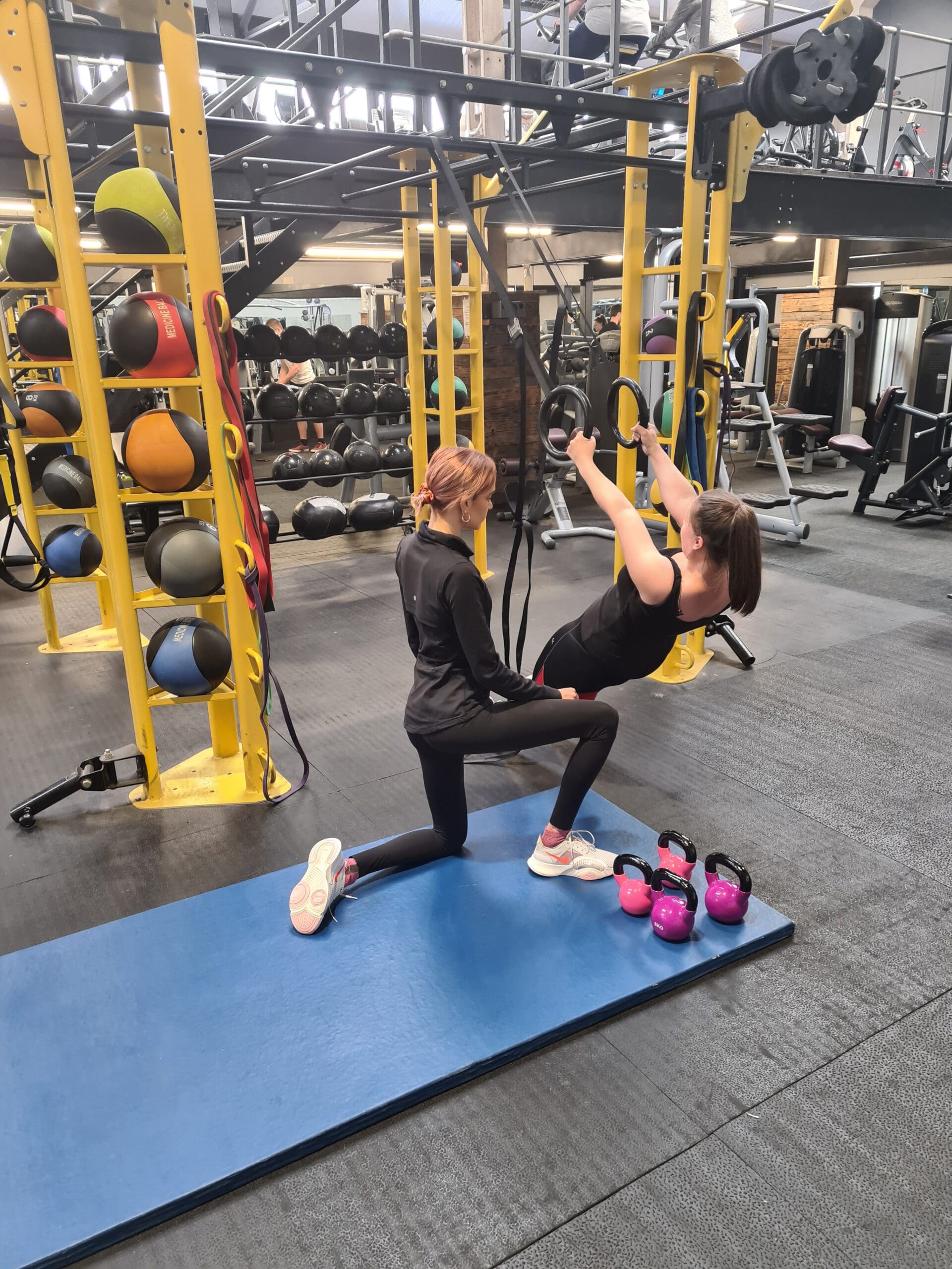 Two women are in a gym. One woman is exercising on rings, supported by another woman kneeling beside her. Kettlebells and colorful medicine balls are in the background, with gym equipment visible throughout the room.
