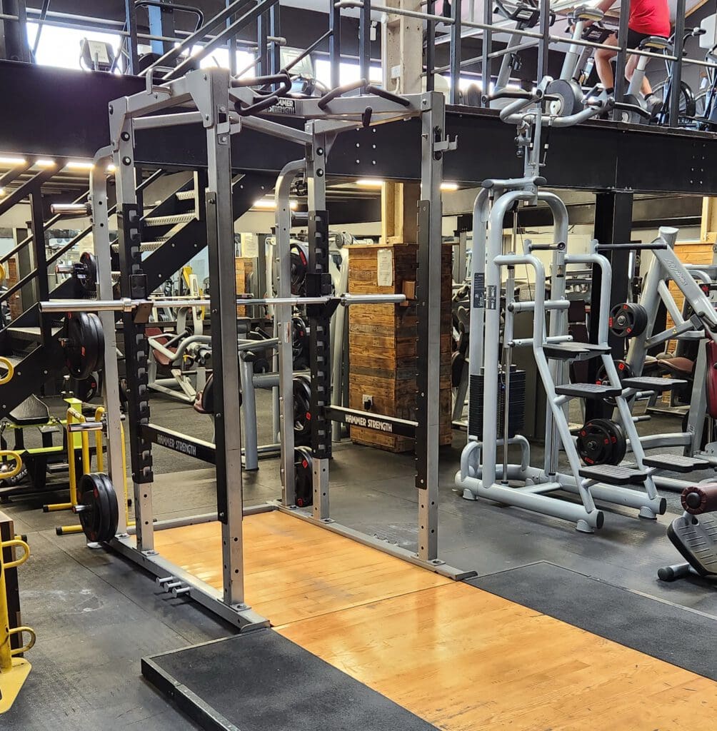 A gym interior featuring a squat rack with weights on a wooden platform. Surrounding equipment includes weight machines and benches. A mezzanine level above shows a person using a cardio machine. The area is well-lit with a mix of metal and wood decor.