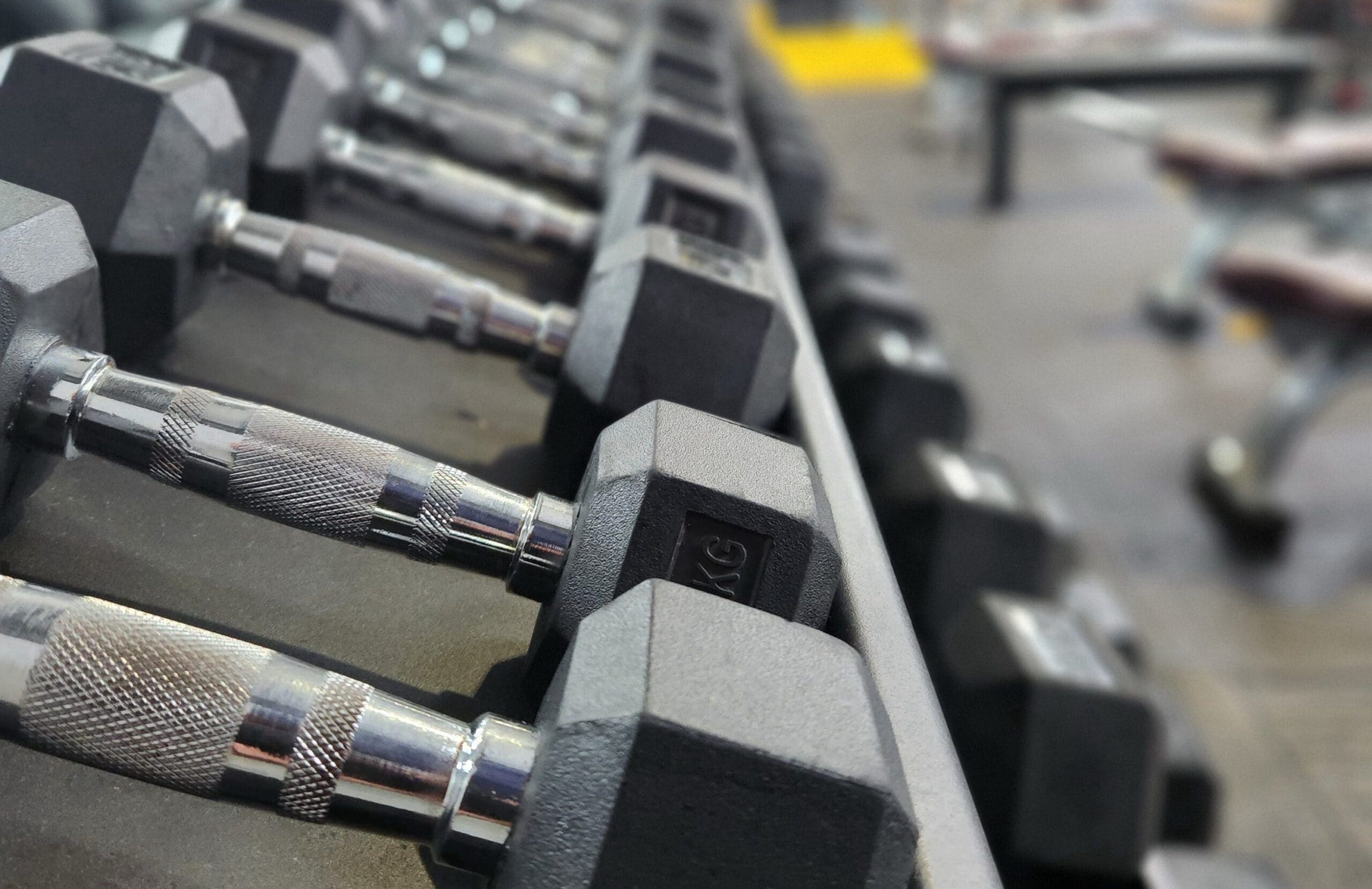 Close-up of a row of hexagonal dumbbells on a rack in a gym, ranging in different weights. The focus is on the nearest dumbbell, with a blurred background of gym equipment and flooring.