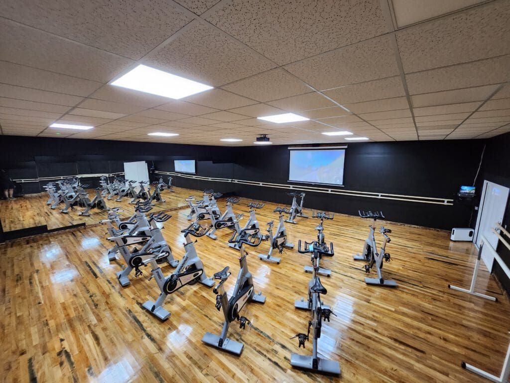 A spacious gym room with numerous stationary bikes arranged in rows on a polished wooden floor. The walls are black with large mirrors and a projector screen at the front. Overhead fluorescent lights illuminate the area.