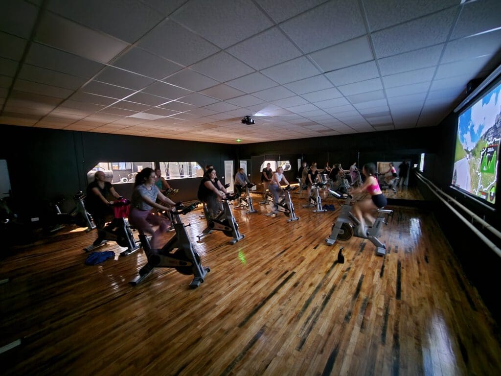 People are participating in an indoor cycling class in a dimly lit room, facing a large screen displaying a virtual landscape. The wooden floor reflects the light, and the cyclists are concentrated on their exercise.