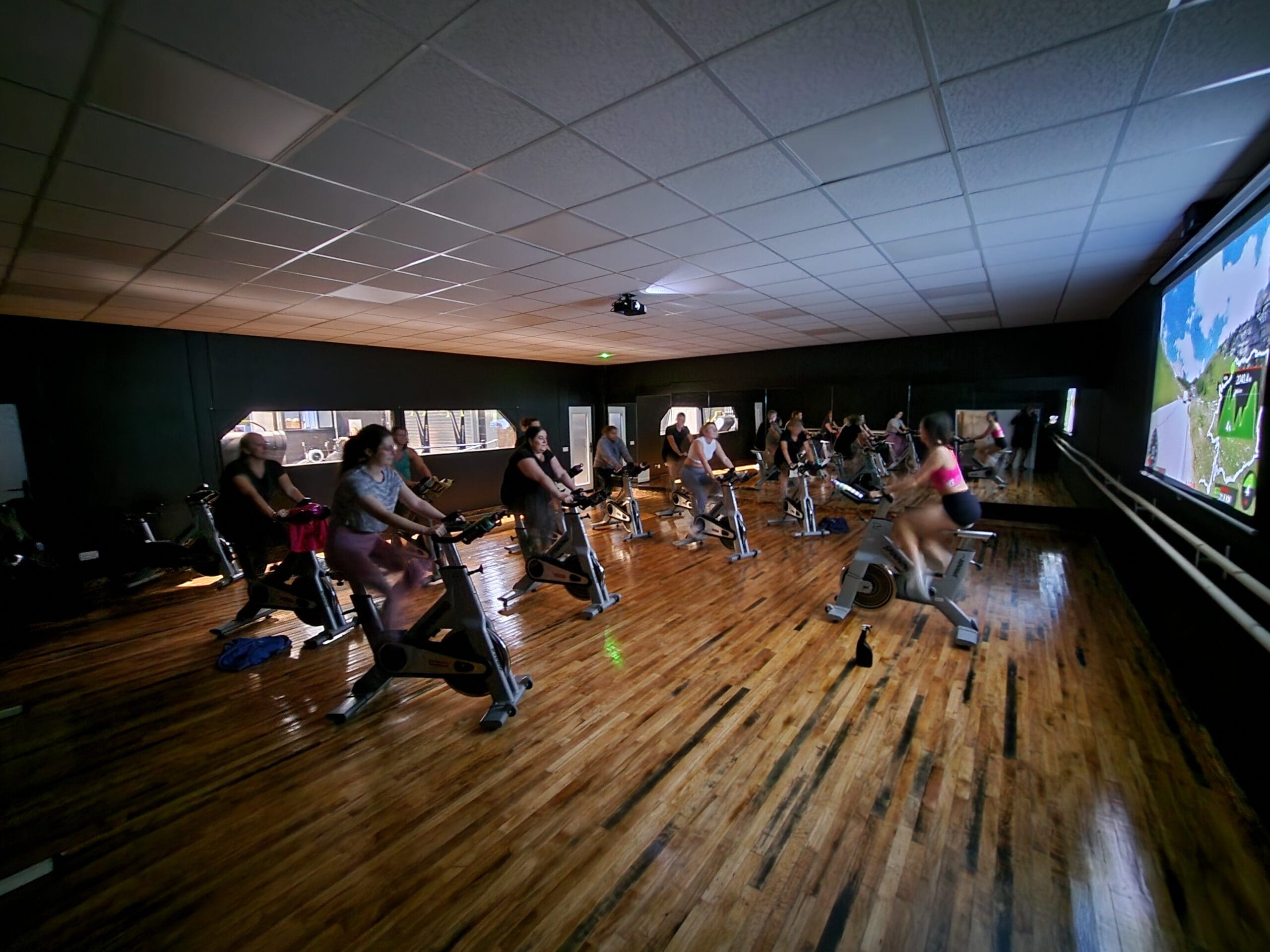 People are participating in an indoor cycling class in a dimly lit room, facing a large screen displaying a virtual landscape. The wooden floor reflects the light, and the cyclists are concentrated on their exercise.