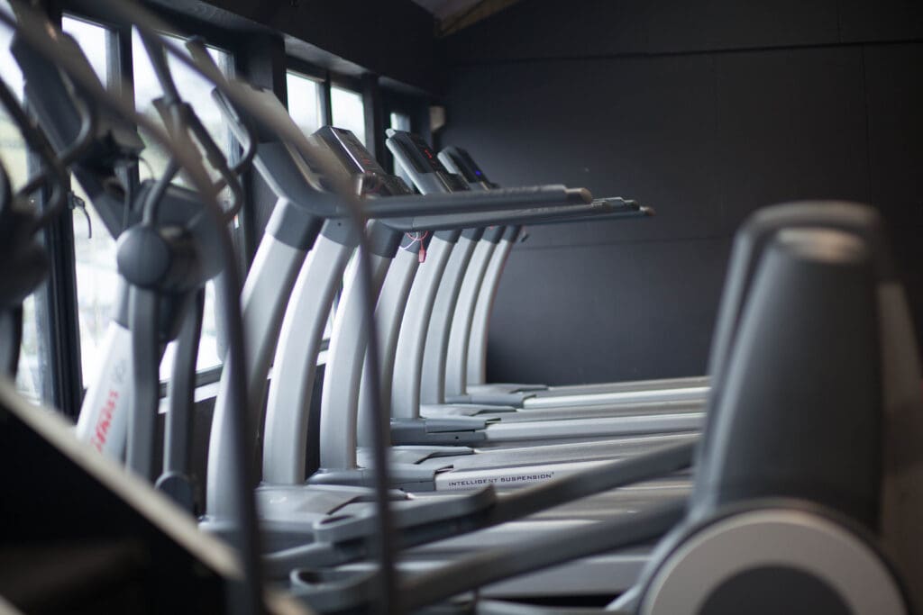 A row of modern treadmills positioned side by side in a gym with large windows on the left, allowing natural light to fill the space. The room has a sleek, minimalist design.