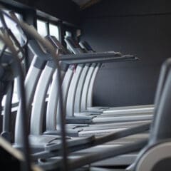 A row of modern treadmills positioned side by side in a gym with large windows on the left, allowing natural light to fill the space. The room has a sleek, minimalist design.