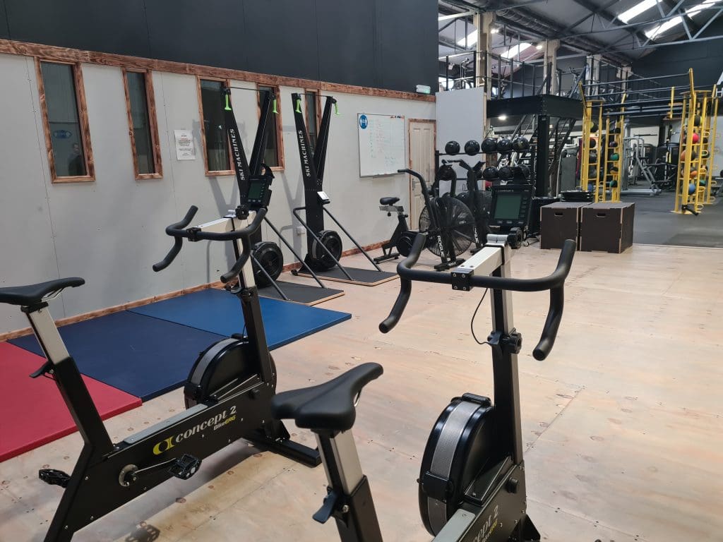 A modern gym interior with exercise bikes in the foreground. In the background, there are rowing machines, a climbing structure, and foam boxes for workouts. The gym has a high ceiling and bright lighting.