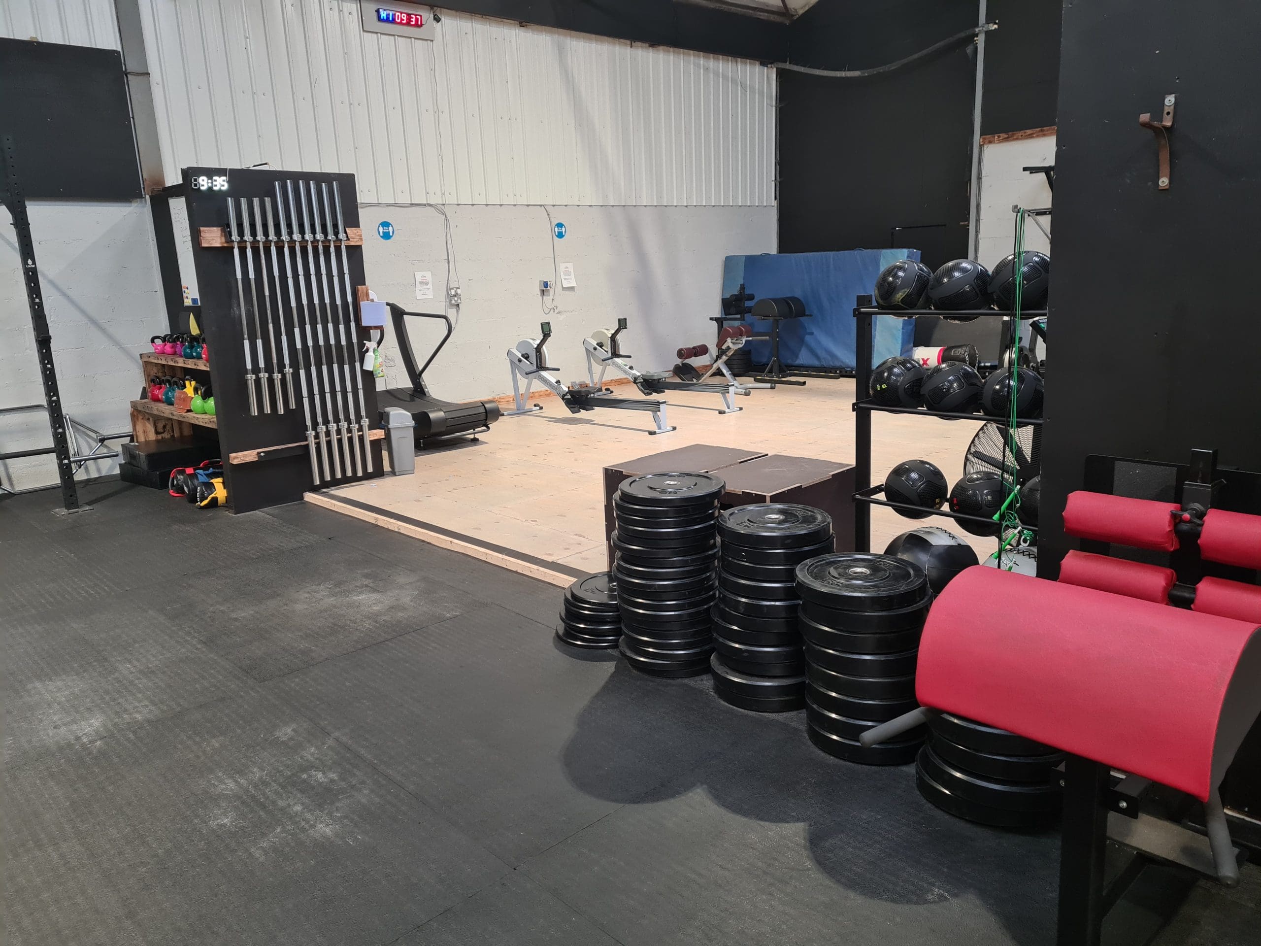 A gym space with exercise bikes, a rowing machine, gymnastic rings, and medicine balls. Weight plates are stacked on the floor, alongside red-padded equipment. The area has a black rack with kettlebells and colorful dumbbells.