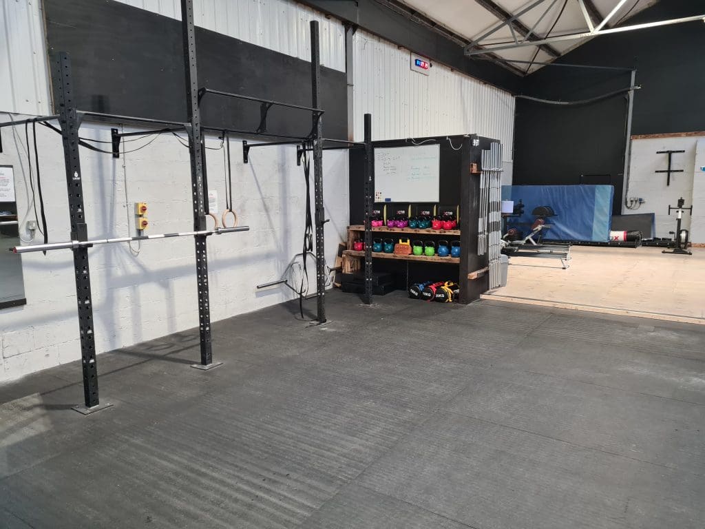 A gym with exercise equipment, including a pull-up bar, rings, kettlebells, and barbells. There's a board with writing, colorful kettlebells on shelves, and a wooden platform on the right. The space has gray mats and a white and black wall.