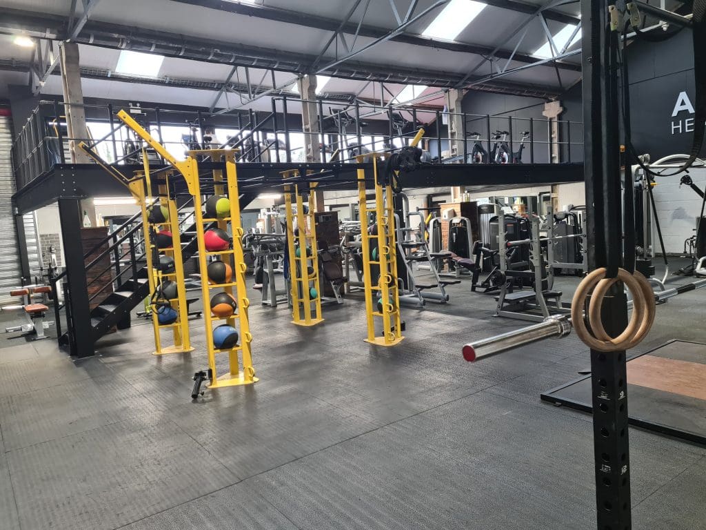 A spacious gym interior with equipment, including a yellow frame with colorful medicine balls, gymnastic rings, weight machines, and cardio equipment. An elevated platform with additional machines is visible in the background.