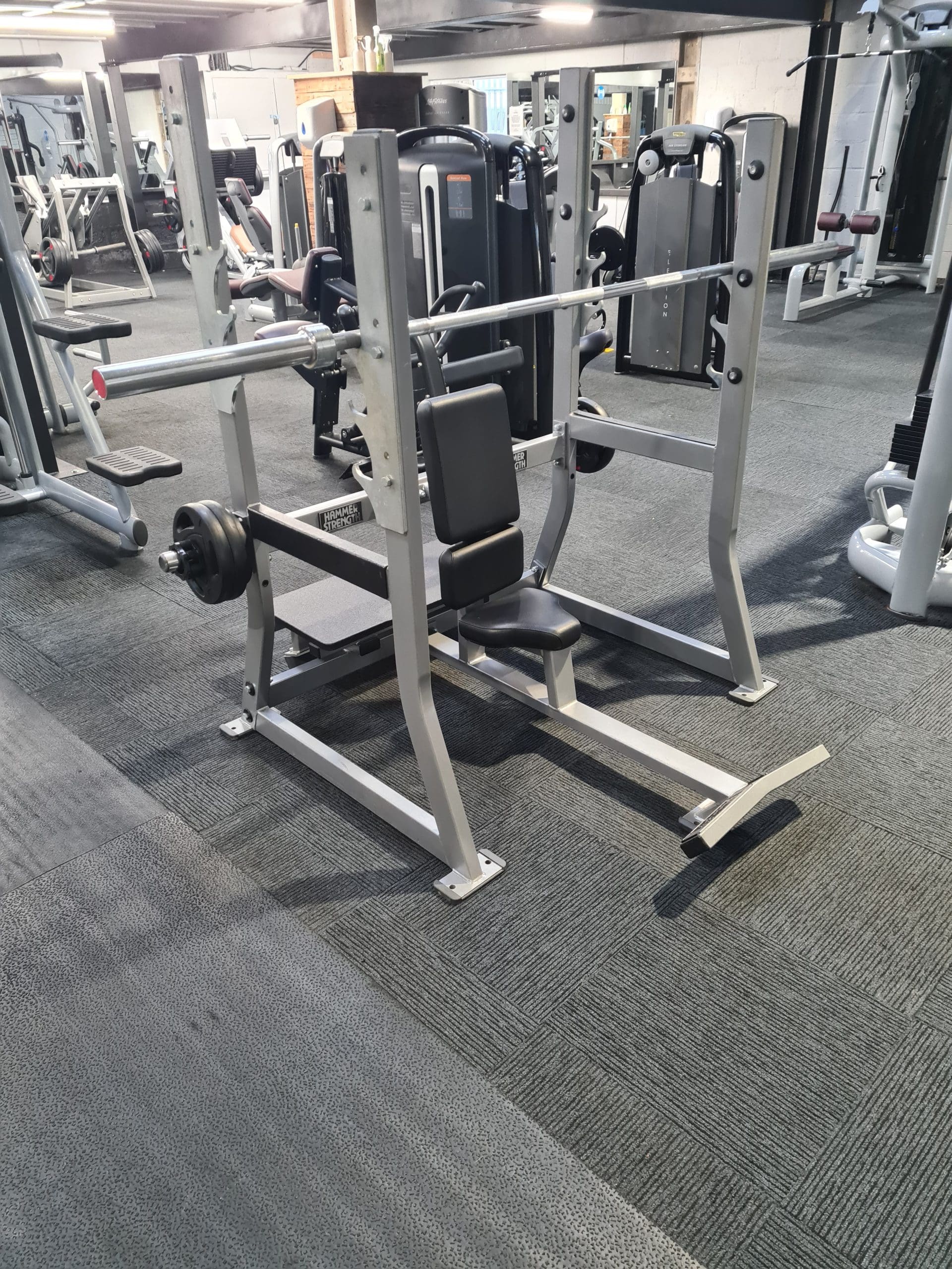 A gym setting with a bench press station featuring a padded bench, barbell rack, and barbells with weights on both ends. Other gym equipment is visible in the background on a carpeted floor.