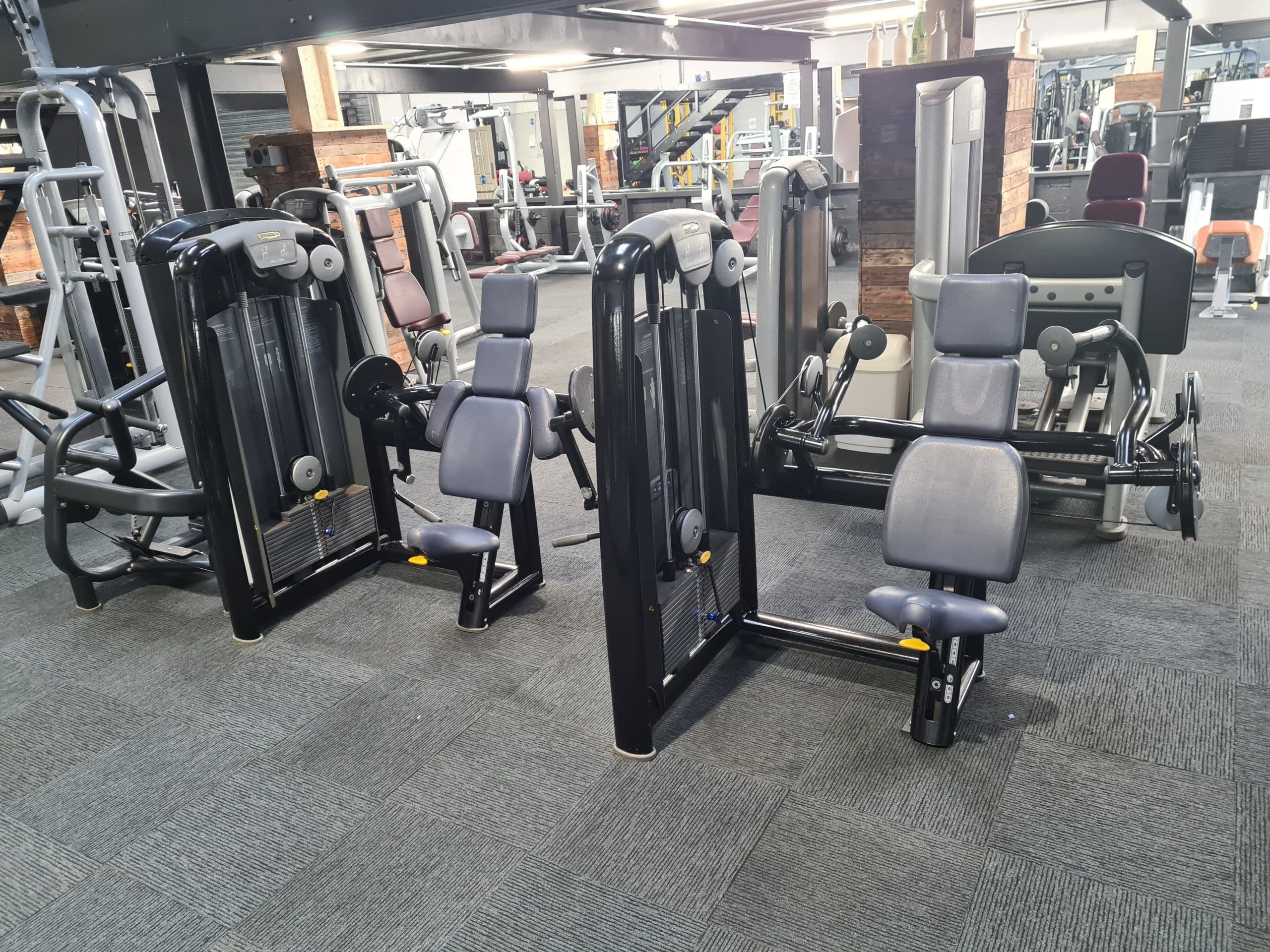 A gym interior showing various exercise machines, including two seated strength machines with padded seats and a leg press machine. The room has gray carpet flooring and is equipped with modern fitness equipment.
