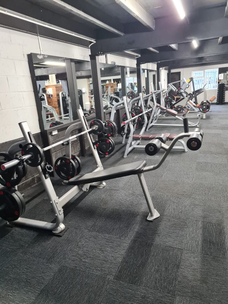 A gym room with various exercise equipment including weightlifting benches, racks filled with barbells, and mirrors on the wall. The floor is carpeted, and the space is well-lit with overhead lights.