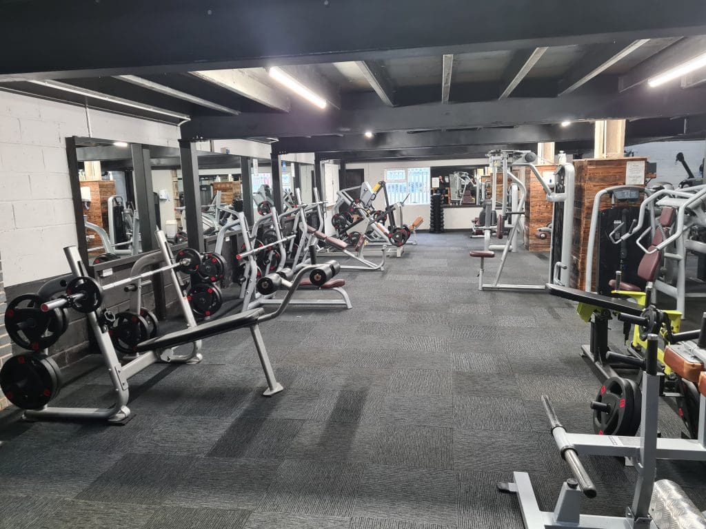 A spacious gym with various weightlifting equipment, including benches, barbells, and machines. The room has a dark, carpeted floor, large mirrors on the walls, and bright overhead lighting, creating an open and inviting workout environment.