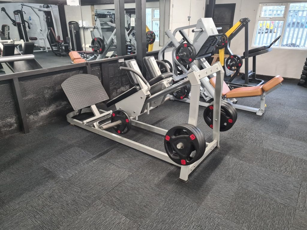 A gym interior featuring a leg press machine with weights loaded on the sides. The machine is set on a gray carpeted floor, surrounded by mirrors and other gym equipment. A nearby window lets in natural light.
