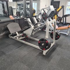 A gym interior featuring a leg press machine with weights loaded on the sides. The machine is set on a gray carpeted floor, surrounded by mirrors and other gym equipment. A nearby window lets in natural light.