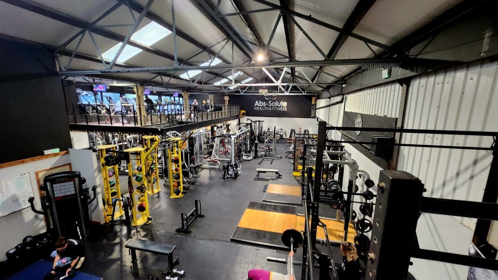 A wide-angle view of a spacious gym with various exercise equipment, including weightlifting machines, free weights, and cardio machines. The gym has high ceilings and a mezzanine level with additional equipment. The floor is mostly black.
