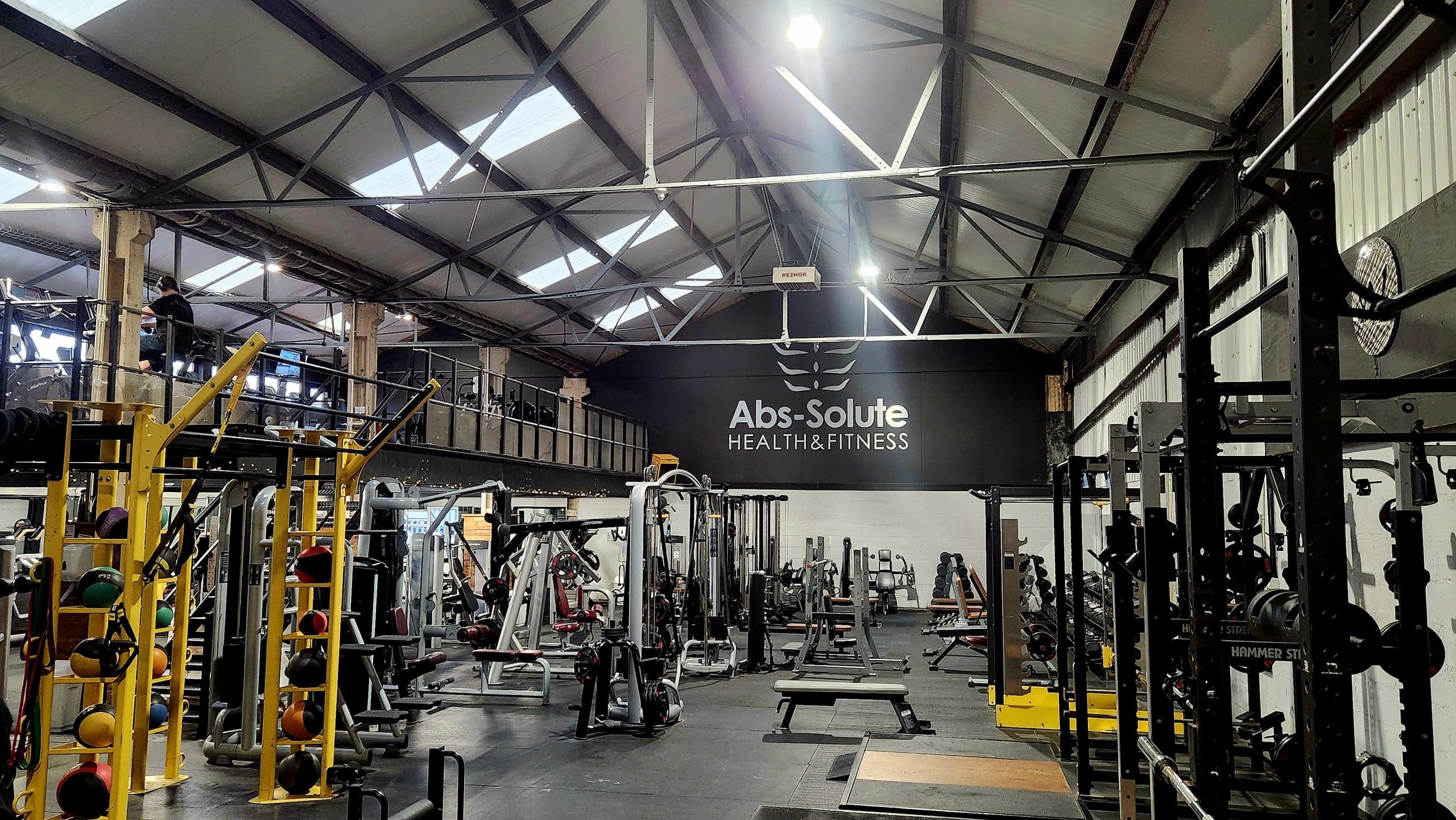 A spacious gym with various exercise equipment, including weight racks, machines, and benches. "Abs-Solute Health & Fitness" is displayed on the wall. The space has high ceilings with skylights and an upper balcony area.