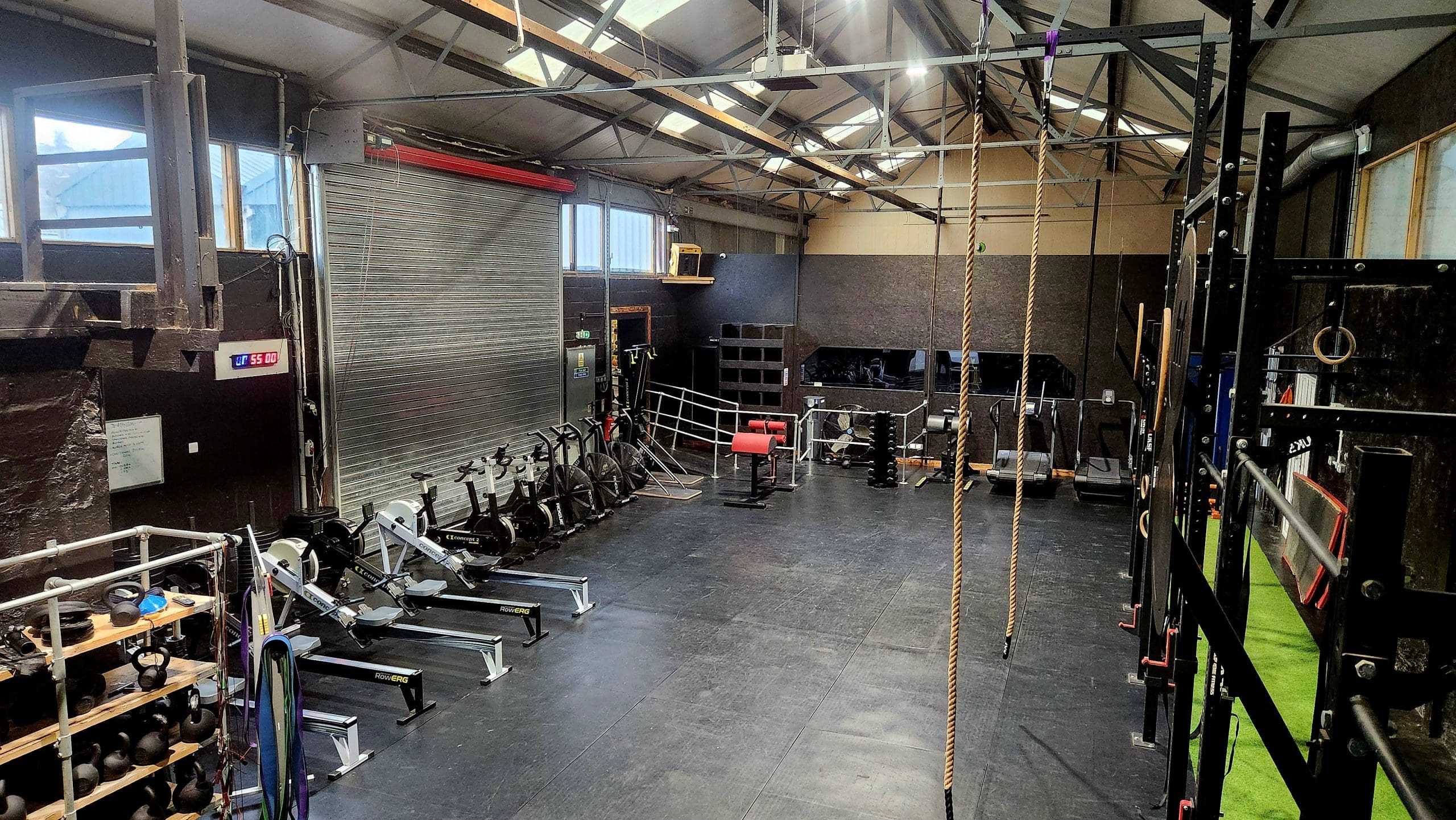 A spacious gym with rowing machines lined up on the left, a climbing rope hanging from the ceiling, a metal staircase at the back, and various fitness equipment along the walls. The floor is black, and large windows let in natural light.