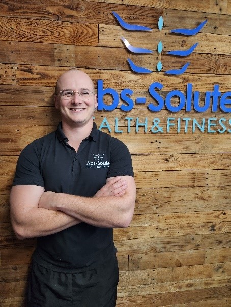 A smiling person with glasses and a shaved head stands with arms crossed in front of a wooden wall. They wear a dark polo shirt with a logo, next to a sign that reads "Abs-Solute Health & Fitness" in blue letters.