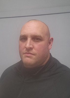 A man with a shaved head and wearing a black zip-up jacket looks at the camera against a plain grey background.