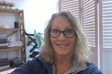 A smiling woman with glasses takes a selfie indoors. She has shoulder-length wavy hair and is wearing a blue zip-up top. Behind her is a wooden shelf with neatly folded towels and a plant, near a window with white shutters.