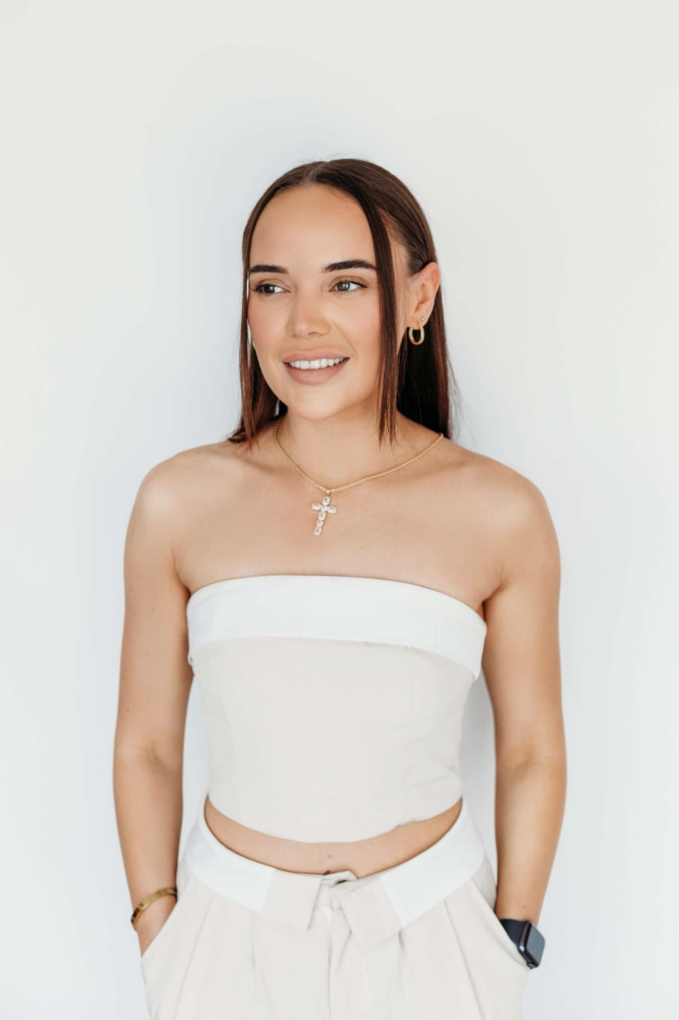 A woman with long brown hair is smiling, wearing a strapless white top and light beige pants. She has a cross necklace, hoop earrings, a bracelet, and a watch. Her hands are in her pockets, standing against a plain white background.