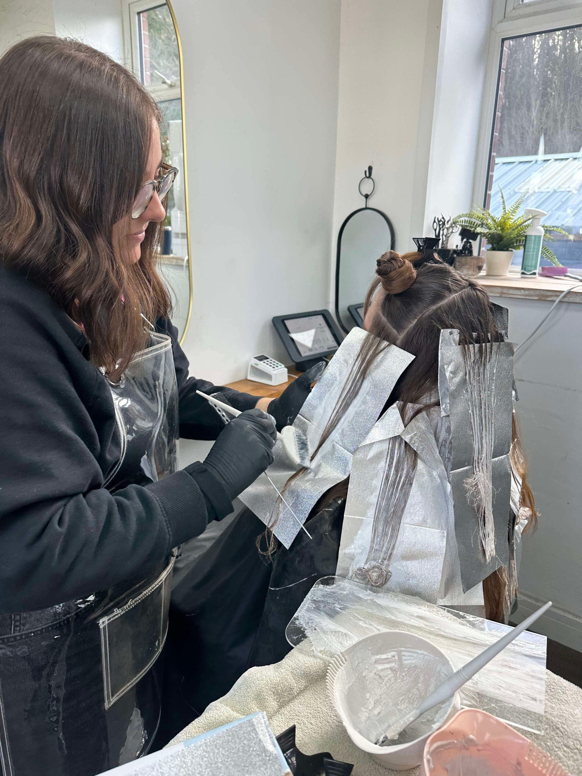 A woman in a salon applies hair dye to another woman's hair using foil. Both are focused on the styling process. The salon workspace includes tools and a window showing outdoor greenery.