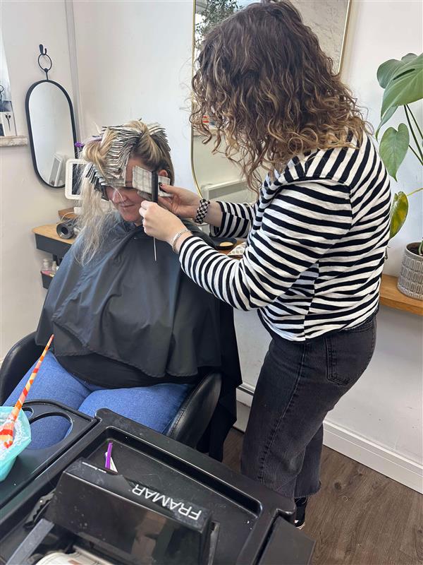 A hairstylist applies hair foils to a client's hair at a salon. The client is seated and wearing a black cape. The hairstylist is wearing a striped shirt. A mirror and a plant are visible in the background.