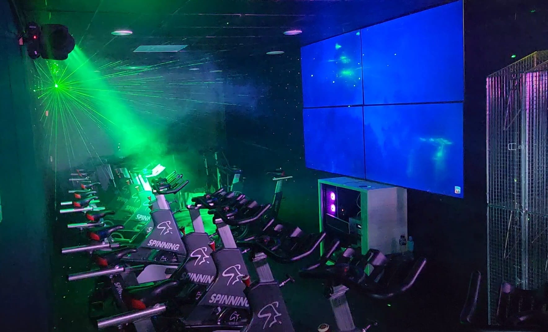 A dimly lit indoor cycling studio features rows of stationary bikes with "Spinning" labels. Laser lights and a large screen displaying abstract blue visuals create an energetic atmosphere.