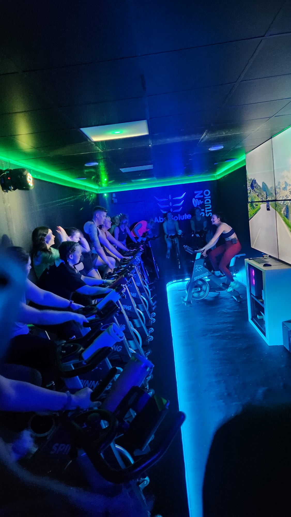 A group exercise class in a dark room with neon lighting. Participants are riding stationary bikes, following an instructor at the front. A large screen displays scenic landscapes, enhancing the immersive experience.